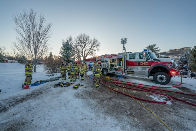 Firefighters in winter gear work near a fire truck in a snowy field.